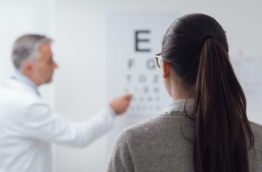 A woman with long hair in a ponytail is undergoing an eye acuity test. A male optician is pointing to a letter on the chart on the wall while the female patient is calling out the letter she sees from a distance.