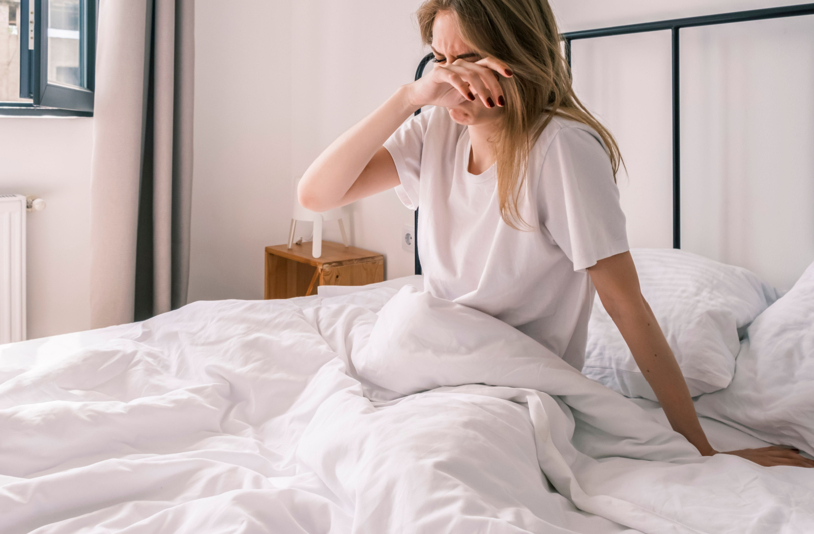 A woman in bed wearing a white shirt rubbing her eyes after waking up.