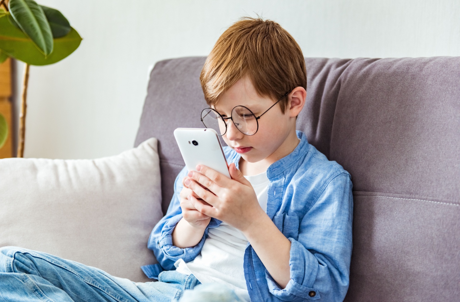 A child with large round glasses sitting on a couch and holding a smartphone very close to his face
