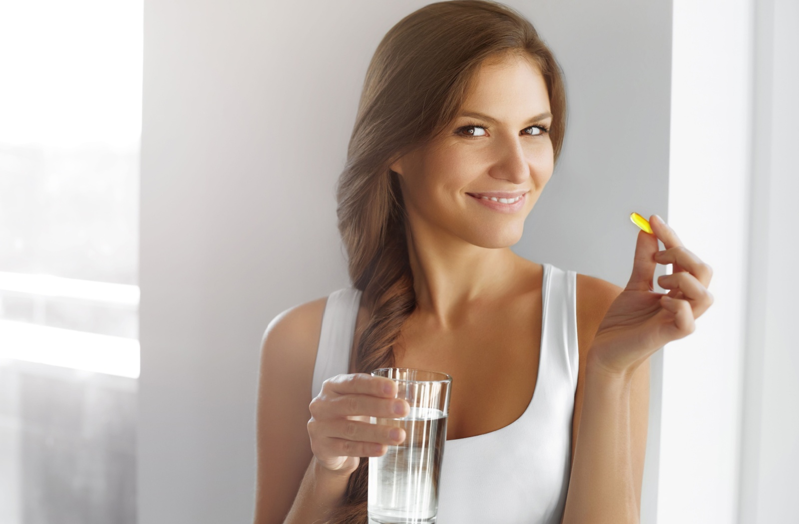 A smiling woman holding a fish oil pill in her left hand and a glass of water in her right hand