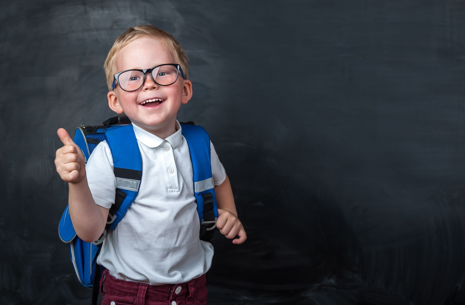 A young child wearing glasses and a blue backpack poses with a thumbs up in front of a grey background