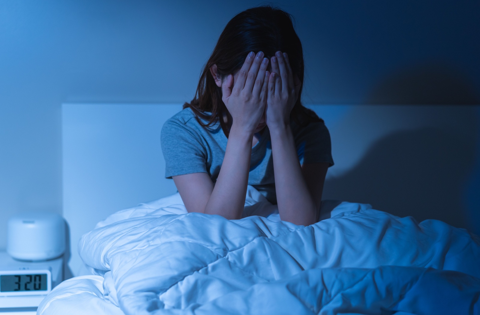 A woman sitting upright in bed in a dark room with both hands over her eyes, suffering from a migraine.