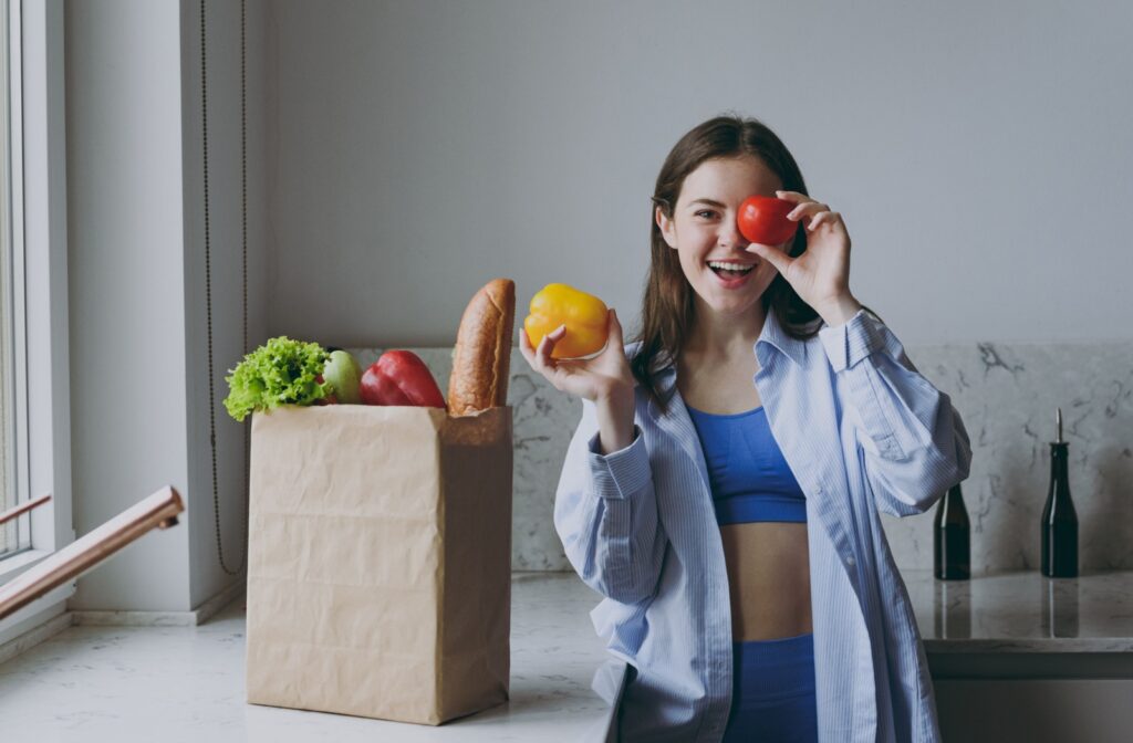 An adult holding fresh vegetables and smiling playfully near a paper bag filled with groceries, including leafy greens, bell peppers, and bread.