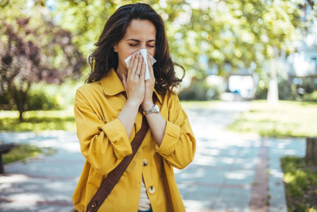 A person with seasonal allergies blows their nose with a tissue while outside.