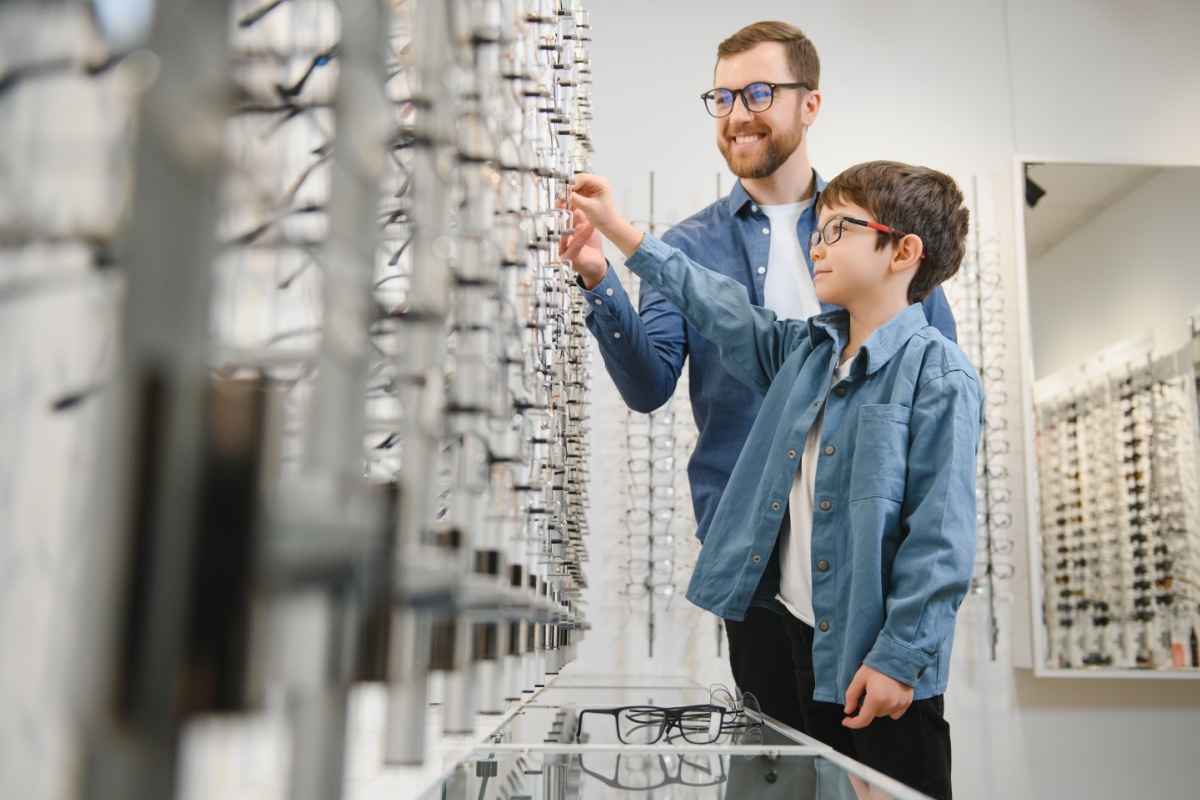 A man and a child browse a wall display of eyeglass frames together inside an optometrist’s office.